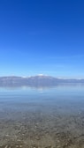 A serene lake scene with crystal-clear water in the foreground, pebbles visible beneath the surface. In the background, snow-capped mountains rise against a vast, cloudless blue sky.
