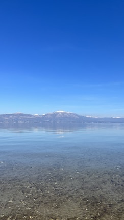A serene lake scene with crystal-clear water in the foreground, pebbles visible beneath the surface. In the background, snow-capped mountains rise against a vast, cloudless blue sky.