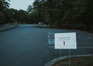 A parking lot with a sign in the foreground indicating pick-up and drop-off for a naturalist camp. The sign has an arrow pointing straight ahead. There are a few cars parked in the distance, and the area is surrounded by dense trees and greenery.