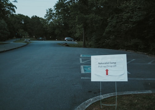 A parking lot with a sign in the foreground indicating pick-up and drop-off for a naturalist camp. The sign has an arrow pointing straight ahead. There are a few cars parked in the distance, and the area is surrounded by dense trees and greenery.