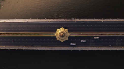 A top-down view of a wide bridge spanning over a body of water. The bridge features several lanes with moving vehicles, and a distinctive decorative circular structure in the center. The water below appears dark and reflects some light, while the bridge is illuminated by golden tones.