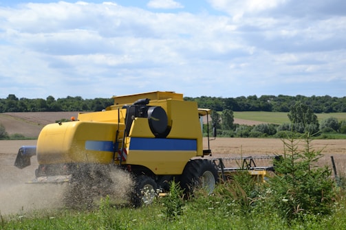 A yellow combine harvester is operating in a field, surrounded by lush greenery and under a partly cloudy sky. The harvester is depicted in motion, cutting through the crop as dust emerges around it.