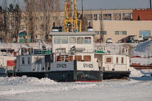 A large boat with identification markings 'BTK-629' is docked on a snowy shore. The scene is set against an industrial backdrop with buildings and parked vehicles. Leafless trees and antennas are visible on the rooftop of the structure behind the boat.