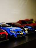 A collection of small model race cars is positioned in a row. The focus is on a vibrant blue race car marked with a Mercedes logo and various decals. Beside it are other cars, some red and black, partially visible and slightly out of focus.