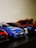 A collection of small model race cars is positioned in a row. The focus is on a vibrant blue race car marked with a Mercedes logo and various decals. Beside it are other cars, some red and black, partially visible and slightly out of focus.