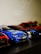 A collection of small model race cars is positioned in a row. The focus is on a vibrant blue race car marked with a Mercedes logo and various decals. Beside it are other cars, some red and black, partially visible and slightly out of focus.