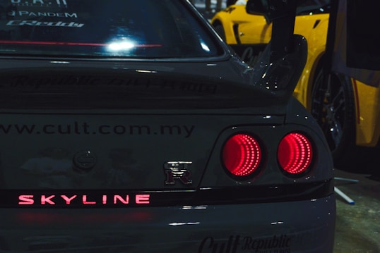 The image features the rear of a black Nissan Skyline R with its distinct circular tail lights illuminated. The car has a prominent spoiler. The text 'Skyline' is lit up in red at the bottom of the trunk. Advertisements for various brands and websites are visible on the rear window and trunk. Additionally, there is a partial view of a yellow sports car to the right.