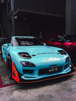 A turquoise sports car with distinctive racing decals and enhancements is parked indoors next to a red car. The car features aerodynamic modifications, including a prominent front splitter with orange accents, and black wheels. The setting is dimly lit, likely a garage or warehouse.