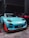 A turquoise sports car with distinctive racing decals and enhancements is parked indoors next to a red car. The car features aerodynamic modifications, including a prominent front splitter with orange accents, and black wheels. The setting is dimly lit, likely a garage or warehouse.