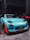 A turquoise sports car with distinctive racing decals and enhancements is parked indoors next to a red car. The car features aerodynamic modifications, including a prominent front splitter with orange accents, and black wheels. The setting is dimly lit, likely a garage or warehouse.