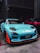 A turquoise sports car with distinctive racing decals and enhancements is parked indoors next to a red car. The car features aerodynamic modifications, including a prominent front splitter with orange accents, and black wheels. The setting is dimly lit, likely a garage or warehouse.