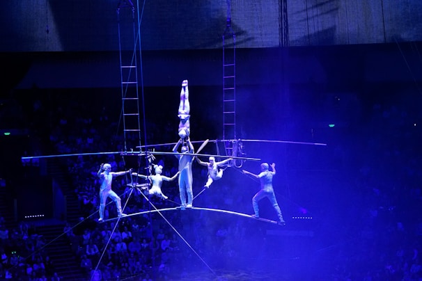 A group of acrobats performs a balancing act on a high wire under dim blue lighting. The performers are engaging in an impressive formation, with one acrobat balancing upside down on another's shoulders while others hold onto a large circular structure. The blue hue creates an atmospheric and dramatic scene within a large, indoor arena with a visible audience.