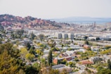 The image provides a wide view of an industrial facility, most likely an oil refinery, situated near a residential area. In the foreground, there are houses, buildings, and green spaces indicative of a suburban neighborhood. The midground displays large industrial buildings and a complex array of pipes and storage tanks. The background features multiple cylindrical storage tanks on a hillside, and a body of water further away.