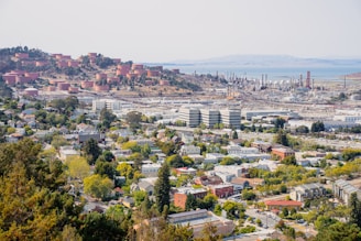 The image provides a wide view of an industrial facility, most likely an oil refinery, situated near a residential area. In the foreground, there are houses, buildings, and green spaces indicative of a suburban neighborhood. The midground displays large industrial buildings and a complex array of pipes and storage tanks. The background features multiple cylindrical storage tanks on a hillside, and a body of water further away.