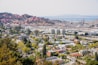 The image provides a wide view of an industrial facility, most likely an oil refinery, situated near a residential area. In the foreground, there are houses, buildings, and green spaces indicative of a suburban neighborhood. The midground displays large industrial buildings and a complex array of pipes and storage tanks. The background features multiple cylindrical storage tanks on a hillside, and a body of water further away.