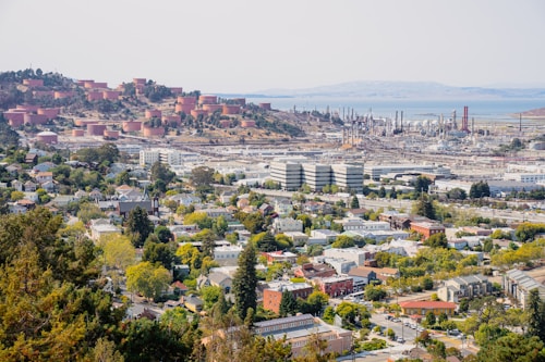 The image provides a wide view of an industrial facility, most likely an oil refinery, situated near a residential area. In the foreground, there are houses, buildings, and green spaces indicative of a suburban neighborhood. The midground displays large industrial buildings and a complex array of pipes and storage tanks. The background features multiple cylindrical storage tanks on a hillside, and a body of water further away.