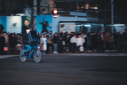 A cyclist rides quickly through a city street at night, carrying a large black parcel on their back. The background is slightly blurred with a crowd of people waiting on the sidewalk and a billboard displaying advertisements. The scene is illuminated by artificial lighting, creating a moody urban atmosphere.