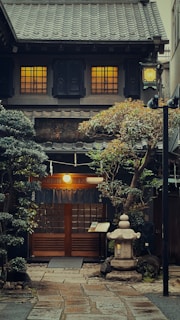 A traditional Japanese building with a tiled roof and illuminated windows. The entrance is framed by blue curtains and surrounded by lush greenery, including neatly trimmed trees and shrubs. A stone lantern and a pathway of stone tiles lead up to the wooden door. The overall ambiance suggests an inviting and serene atmosphere.