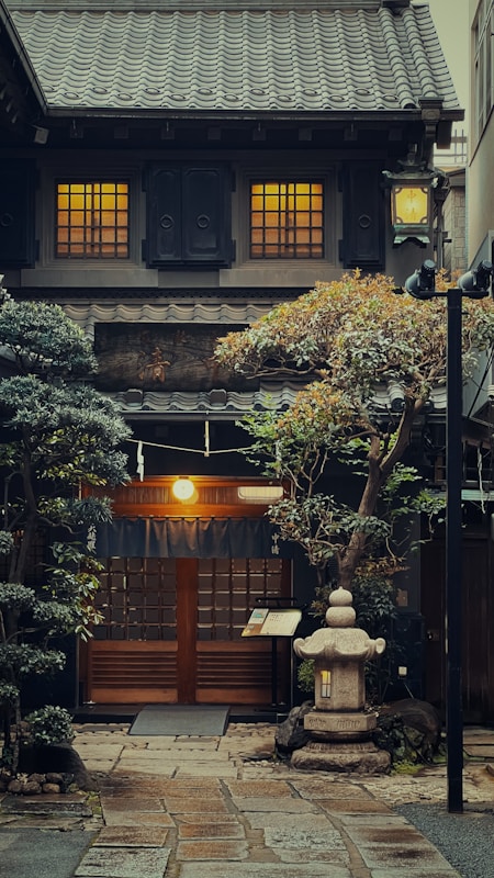A traditional Japanese building with a tiled roof and illuminated windows. The entrance is framed by blue curtains and surrounded by lush greenery, including neatly trimmed trees and shrubs. A stone lantern and a pathway of stone tiles lead up to the wooden door. The overall ambiance suggests an inviting and serene atmosphere.