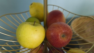 A fruit basket with a variety of fruits including a green apple, a red apple, a lemon, and a pear is neatly arranged in an elegant gold-wire basket against a soft teal background.