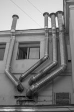 Several large metal ventilation pipes protrude from the side of a building. The pipes are arranged in a geometric pattern and extend vertically upward. The building facade is plain and appears slightly worn, with rectangular windows visible on the left side.