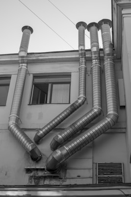 Several large metal ventilation pipes protrude from the side of a building. The pipes are arranged in a geometric pattern and extend vertically upward. The building facade is plain and appears slightly worn, with rectangular windows visible on the left side.