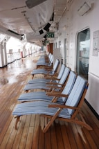 A row of blue-striped lounge chairs is neatly arranged on a wooden deck. The deck is part of a ship, with lifeboats hanging overhead. Signs and emergency instructions are visible on the walls, and there are reflective, glass doors along the side.
