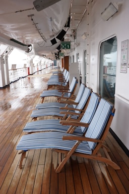 A row of blue-striped lounge chairs is neatly arranged on a wooden deck. The deck is part of a ship, with lifeboats hanging overhead. Signs and emergency instructions are visible on the walls, and there are reflective, glass doors along the side.
