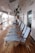 A row of blue-striped lounge chairs is neatly arranged on a wooden deck. The deck is part of a ship, with lifeboats hanging overhead. Signs and emergency instructions are visible on the walls, and there are reflective, glass doors along the side.