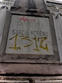 A weathered concrete sign on a building facade with engraved and spray-painted text. The engraved text includes 'CARRETERA GARCIA MORENO', while the spray-painted text in red and yellow partly covers the sign. The structure features classical architectural elements.