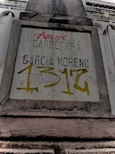 A weathered concrete sign on a building facade with engraved and spray-painted text. The engraved text includes 'CARRETERA GARCIA MORENO', while the spray-painted text in red and yellow partly covers the sign. The structure features classical architectural elements.