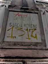 A weathered concrete sign on a building facade with engraved and spray-painted text. The engraved text includes 'CARRETERA GARCIA MORENO', while the spray-painted text in red and yellow partly covers the sign. The structure features classical architectural elements.