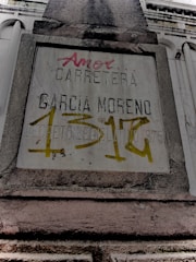 A weathered concrete sign on a building facade with engraved and spray-painted text. The engraved text includes 'CARRETERA GARCIA MORENO', while the spray-painted text in red and yellow partly covers the sign. The structure features classical architectural elements.
