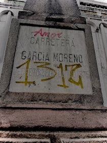 A weathered concrete sign on a building facade with engraved and spray-painted text. The engraved text includes 'CARRETERA GARCIA MORENO', while the spray-painted text in red and yellow partly covers the sign. The structure features classical architectural elements.