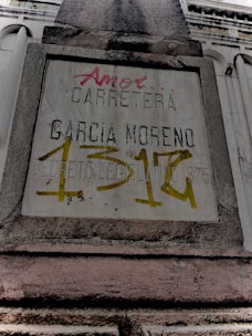 A weathered concrete sign on a building facade with engraved and spray-painted text. The engraved text includes 'CARRETERA GARCIA MORENO', while the spray-painted text in red and yellow partly covers the sign. The structure features classical architectural elements.