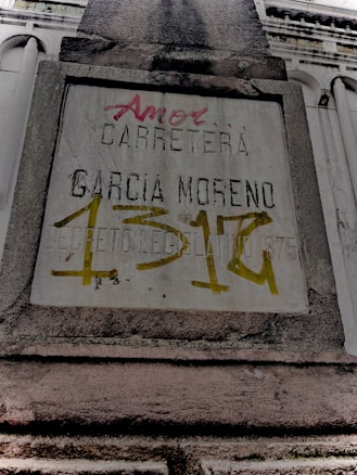 A weathered concrete sign on a building facade with engraved and spray-painted text. The engraved text includes 'CARRETERA GARCIA MORENO', while the spray-painted text in red and yellow partly covers the sign. The structure features classical architectural elements.