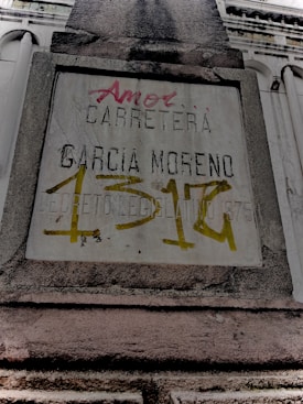 A weathered concrete sign on a building facade with engraved and spray-painted text. The engraved text includes 'CARRETERA GARCIA MORENO', while the spray-painted text in red and yellow partly covers the sign. The structure features classical architectural elements.