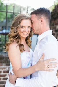 A couple embraces outdoors, with a woman smiling directly at the camera while a man rests his forehead against her temple. They are dressed in light clothing, and the background features a blurred green and brown mix of nature and a brick wall, creating a soft and romantic setting.