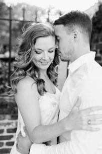 A couple shares a tender moment, embracing closely with expressions of affection and contentment. The image is in black and white, highlighting the couple's soft facial expressions and intimate posture standing near a brick wall with a blurred background.