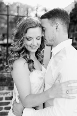 A couple shares a tender moment, embracing closely with expressions of affection and contentment. The image is in black and white, highlighting the couple's soft facial expressions and intimate posture standing near a brick wall with a blurred background.