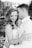 A couple shares a tender moment, embracing closely with expressions of affection and contentment. The image is in black and white, highlighting the couple's soft facial expressions and intimate posture standing near a brick wall with a blurred background.