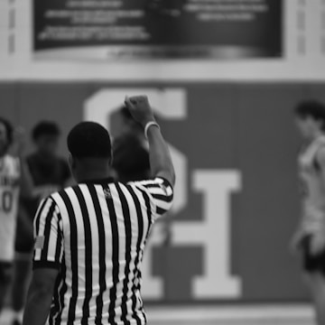 A basketball referee is seen from behind, raising an arm with a closed fist, wearing a black and white striped shirt. The setting appears to be indoors with blurred individuals and a large letter 'S' in the background.