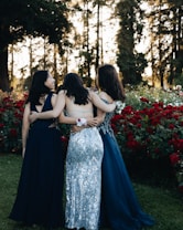 Three women in formal dresses are standing together in a garden, surrounded by vibrant red roses and tall trees in the background. Each woman is wearing a different dress, featuring intricate patterns and designs. They are embracing each other from behind, creating a sense of camaraderie.