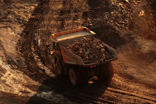 A large dump truck is carrying a load of rocks and soil on a dirt path in a mining or construction site. The surroundings are dusty with tracks from heavy machinery visible on the ground.
