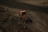 A large yellow bulldozer is positioned on a dirt site, surrounded by loose soil and rocks. Tire tracks are visible in the ground, indicating frequent usage of the area for construction or excavation.