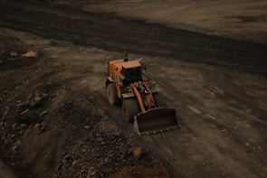 A large yellow bulldozer is positioned on a dirt site, surrounded by loose soil and rocks. Tire tracks are visible in the ground, indicating frequent usage of the area for construction or excavation.