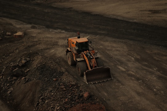 A large yellow bulldozer is positioned on a dirt site, surrounded by loose soil and rocks. Tire tracks are visible in the ground, indicating frequent usage of the area for construction or excavation.