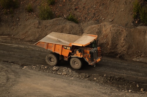 A large industrial dump truck is moving along a dirt road surrounded by rocky terrain. The truck has a robust, orange body with heavy-duty tires suitable for off-road conditions.