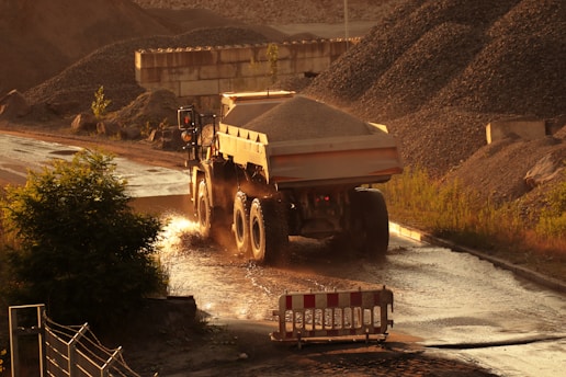 An industrial scene with a large dump truck carrying gravel driving along a dusty road. The surroundings include piles of gravel, patches of vegetation, and a barrier on the road. The lighting suggests a late afternoon or early evening setting with warm tones.