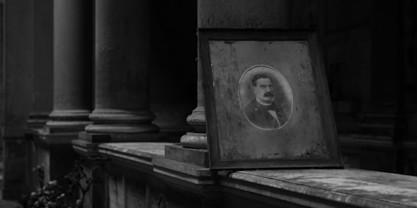 A black and white photograph of a vintage portrait encased in a wooden frame, leaning against the base of a row of large stone columns. The atmosphere appears solemn and historical.